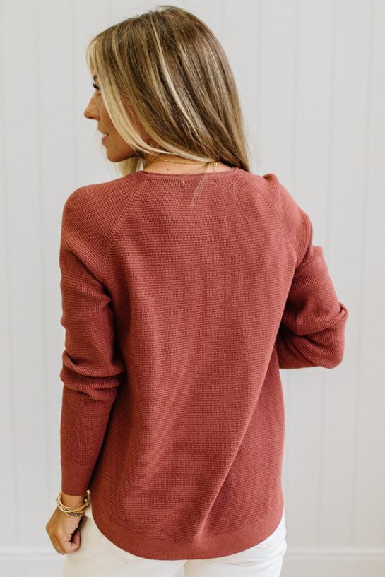 A woman shown from the back wearing a textured, ribbed knit long-sleeve sweater in a dusty rose or terracotta color, paired with white pants against a white paneled wall.