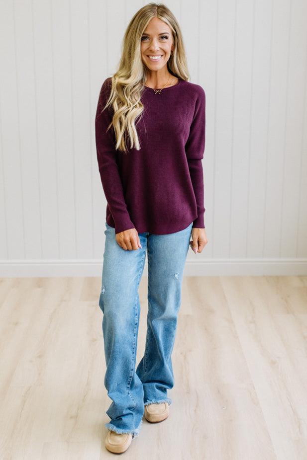 A woman with blonde hair stands in a room with white paneled walls and light wood floors, wearing a long-sleeve crewneck sweater in deep burgundy and light-wash blue jeans.