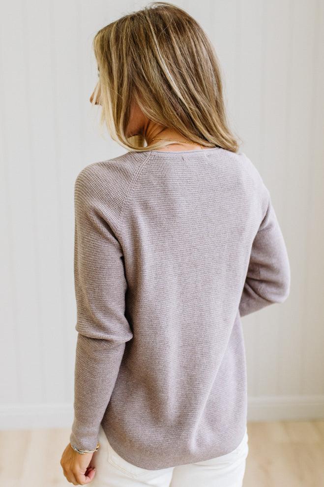 A woman with blonde hair is shown from behind wearing a light taupe ribbed knit sweater and white pants, standing against a white paneled wall.