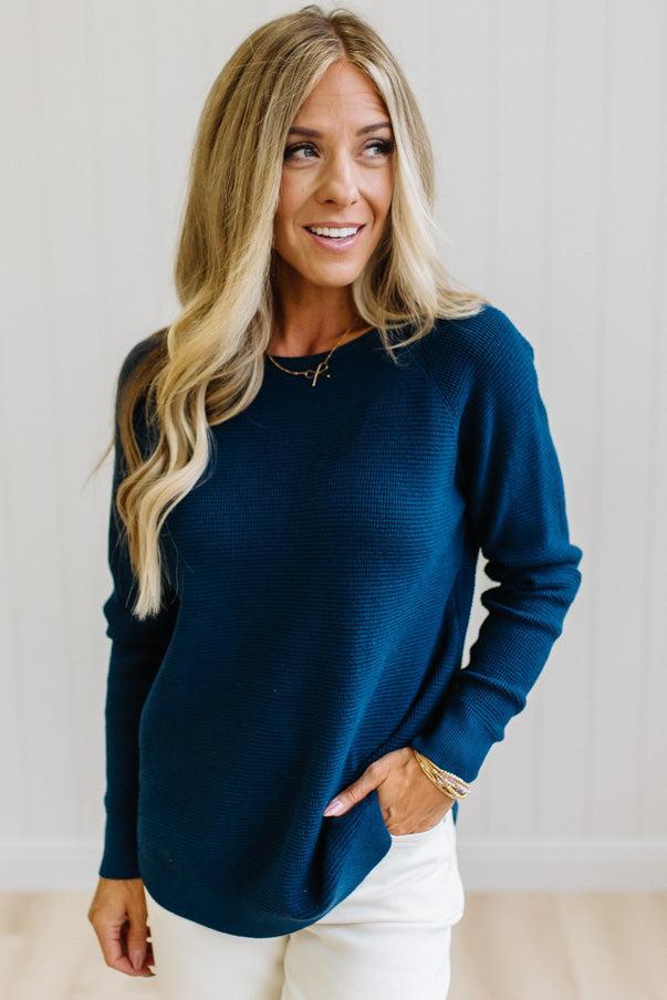 A woman with blonde hair wearing a dark navy blue ribbed knit long-sleeve top and white pants, standing against a white paneled wall.