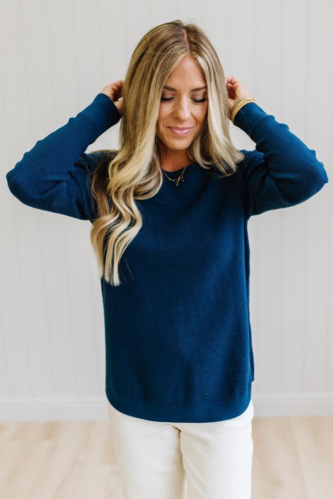 A woman with blonde hair wearing a dark navy blue long-sleeve knit sweater and white pants, standing against a white paneled wall.