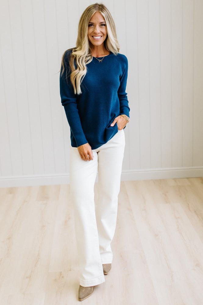 A woman modeling a dark navy blue long-sleeve knit top and white wide-leg pants, standing in a bright room with light wood floors and white paneled walls.