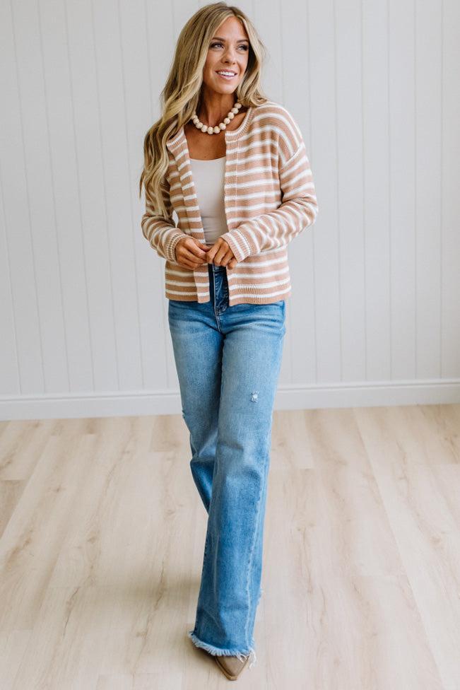 A woman modeling a beige and white striped cardigan over a white tank top and blue jeans, standing in a bright room with light wood floors and white paneled walls.