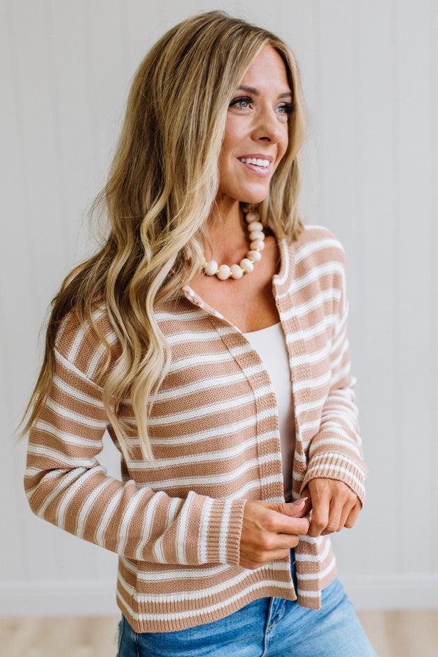 A woman with long blonde hair wearing a beige and white striped cardigan over a white top and blue jeans, accessorized with a cream beaded necklace, posing against a light gray paneled wall.