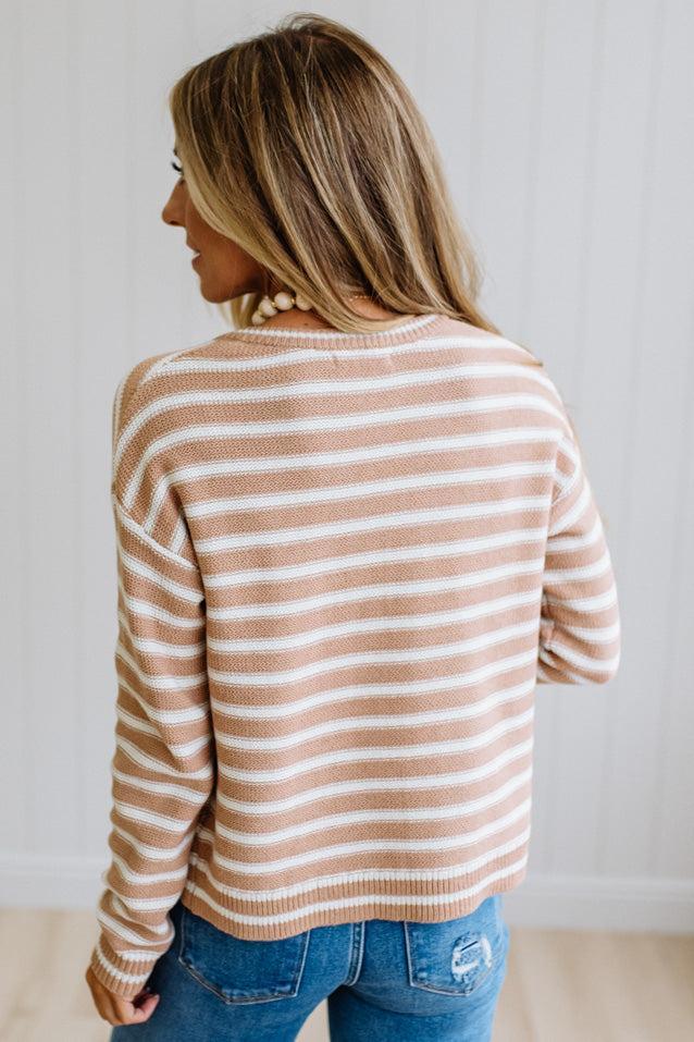 A woman shown from the back wearing a beige and white horizontally striped knit sweater with blue jeans, standing against a white paneled wall.