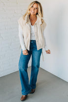 Woman wearing a beige vest, white shirt, blue jeans, and brown shoes against a white brick wall.