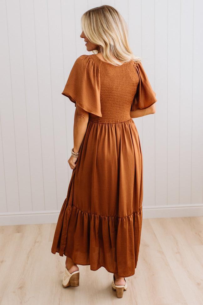 A woman wearing a rust-brown tiered maxi dress with flutter sleeves, shown from the back against a white paneled wall and light wood floor.
