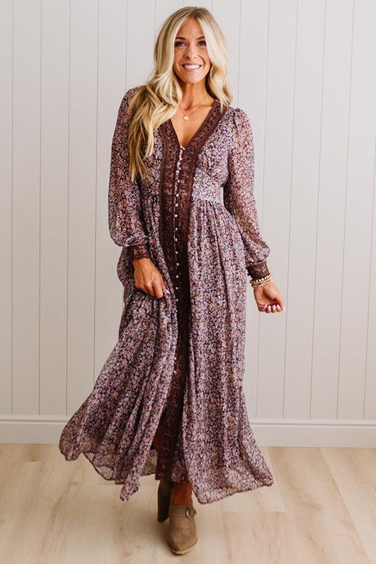 A woman modeling a long-sleeve, V-neck maxi dress with a brown and purple floral pattern, standing in front of a white paneled wall and light wood floor.