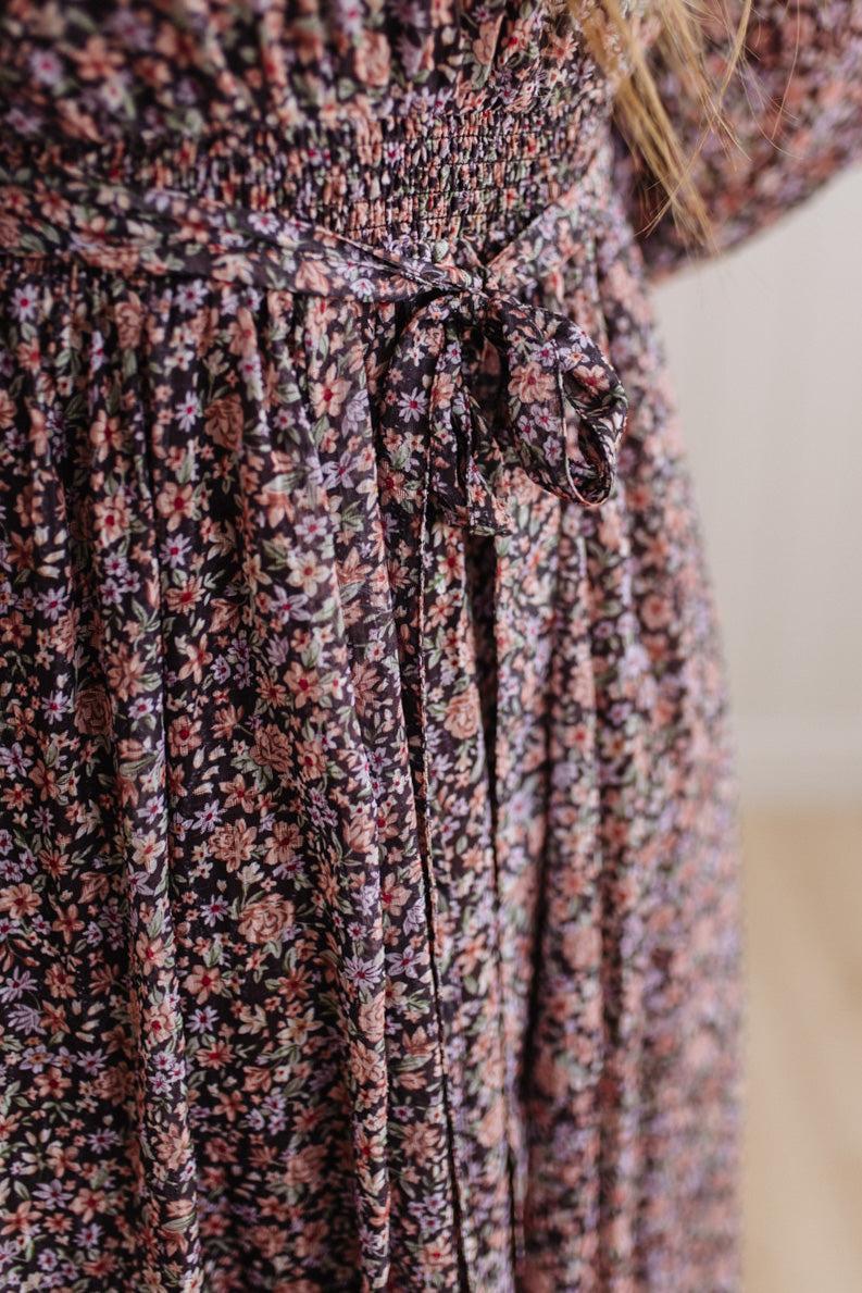 Close-up of a smocked floral dress with a tie waist, featuring a dense pattern of pink, purple, and white flowers on a dark background.