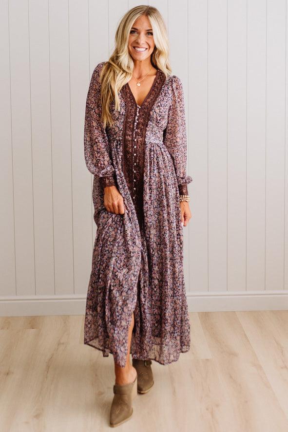 A woman modeling a long-sleeved, button-front maxi dress with a dense floral pattern in dusty purple, brown, and cream, paired with brown suede ankle boots, standing in front of a white paneled wall and light wood floor.