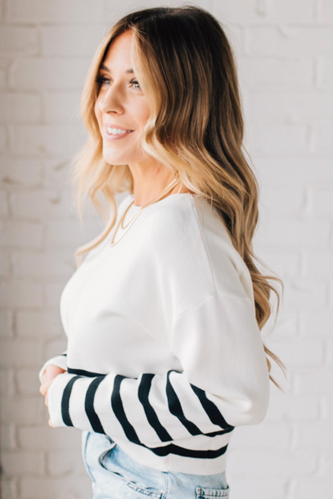 Blonde woman modeling an ivory waist length sweater with black stripes on the bottom half