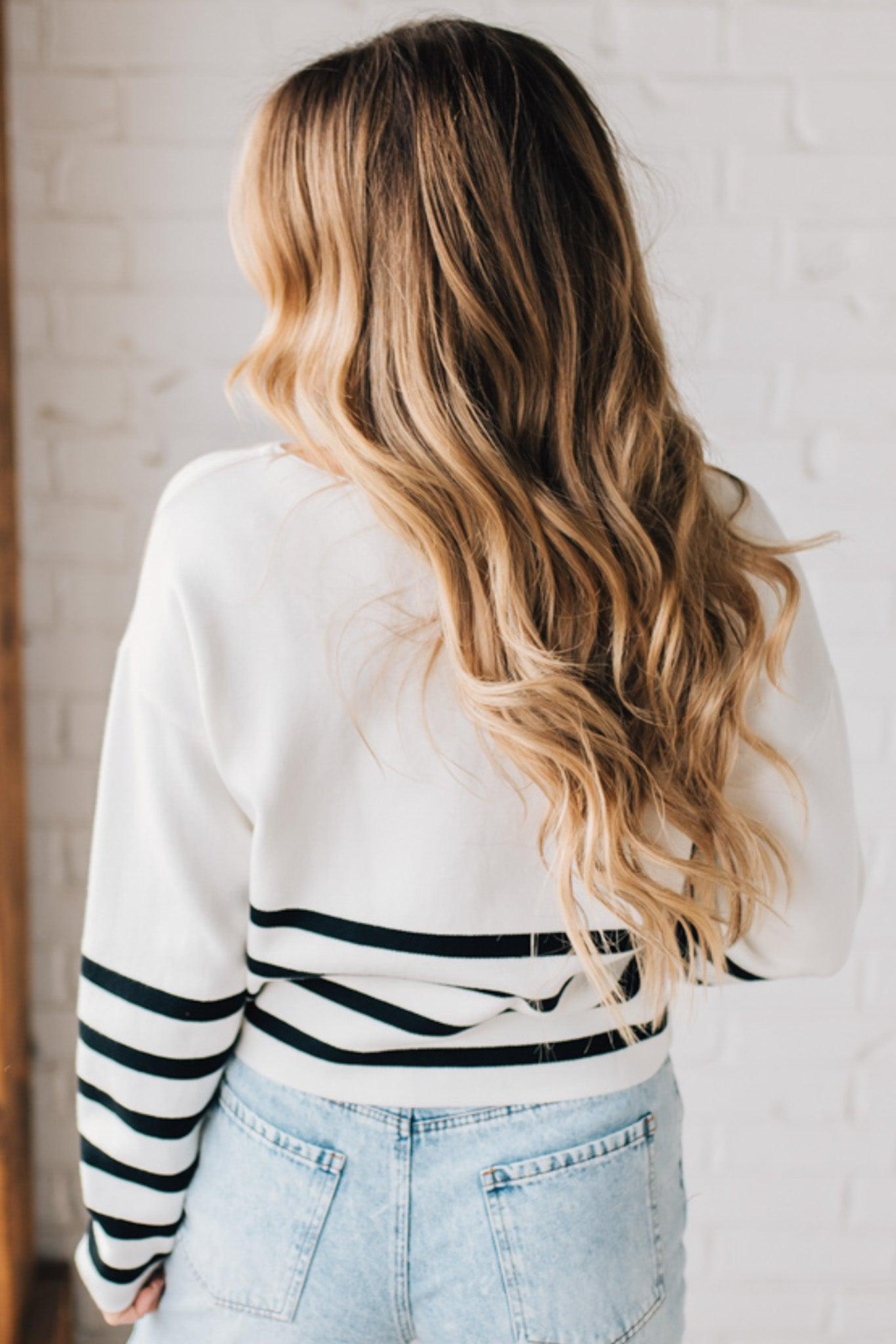 Blonde woman modeling an ivory waist length sweater with black stripes on the bottom half