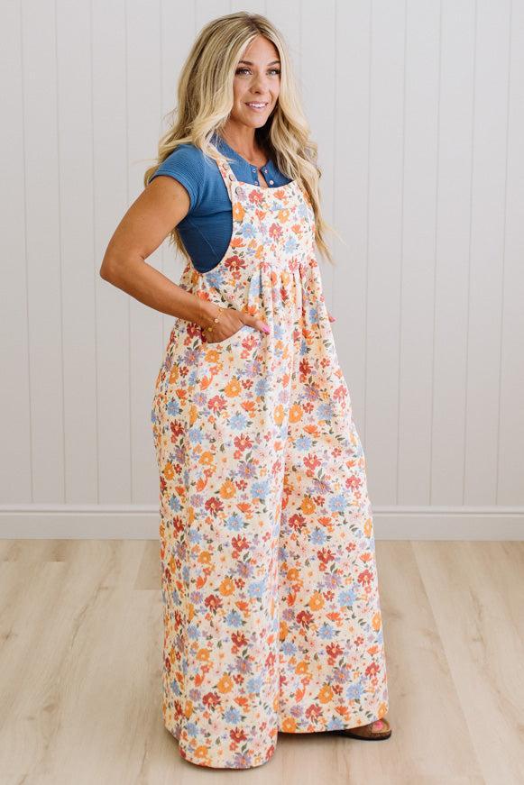 Woman wearing a floral wide overall set over a blue shirt against a white wall.