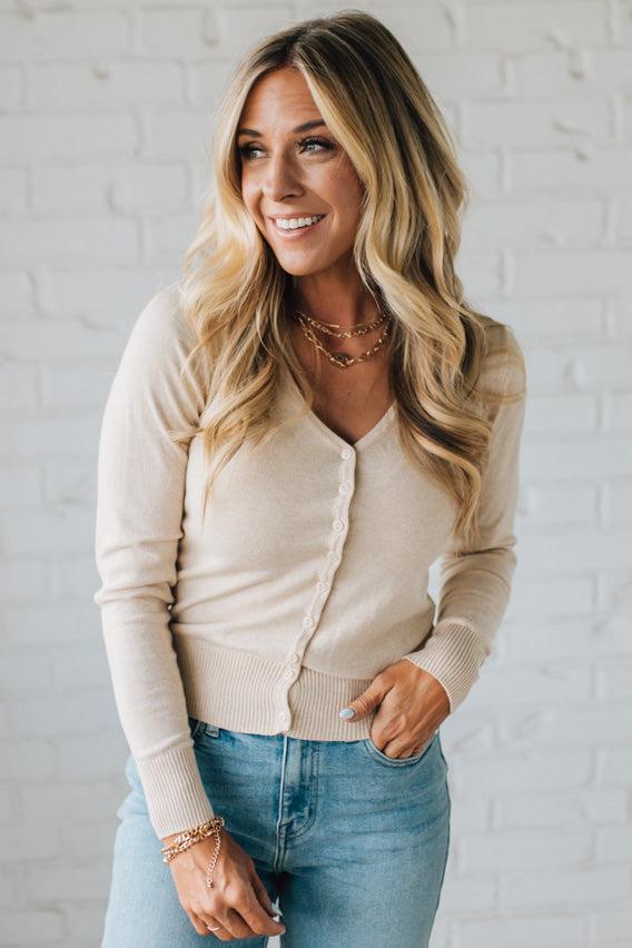 Woman wearing a beige cardigan and blue jeans against a white brick wall.