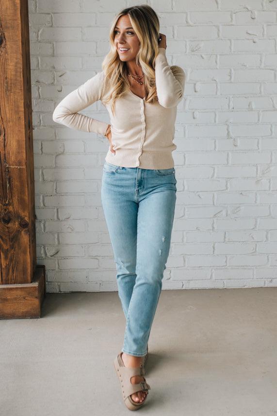 Woman wearing a beige cardigan and light blue jeans against a white brick wall.