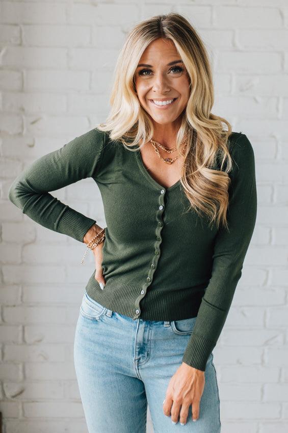 Woman wearing a green long-sleeve top and light blue jeans against a white brick wall.