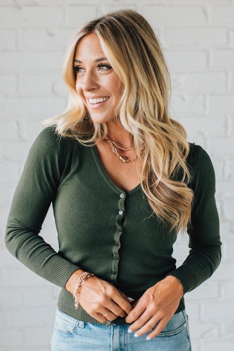 Woman wearing a green cardigan and blue jeans against a white brick wall.