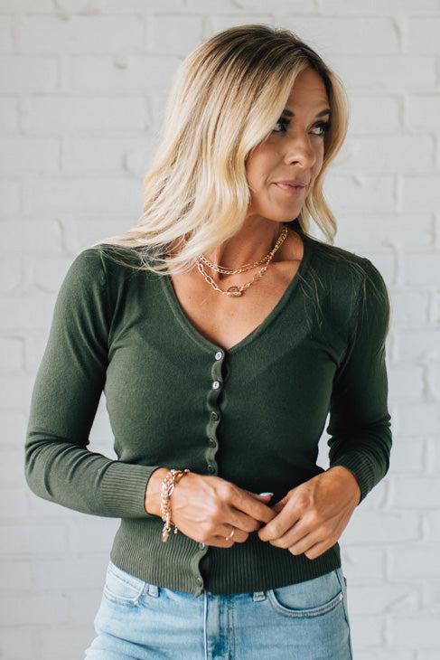 Woman wearing a green cardigan and blue jeans against a white brick wall.
