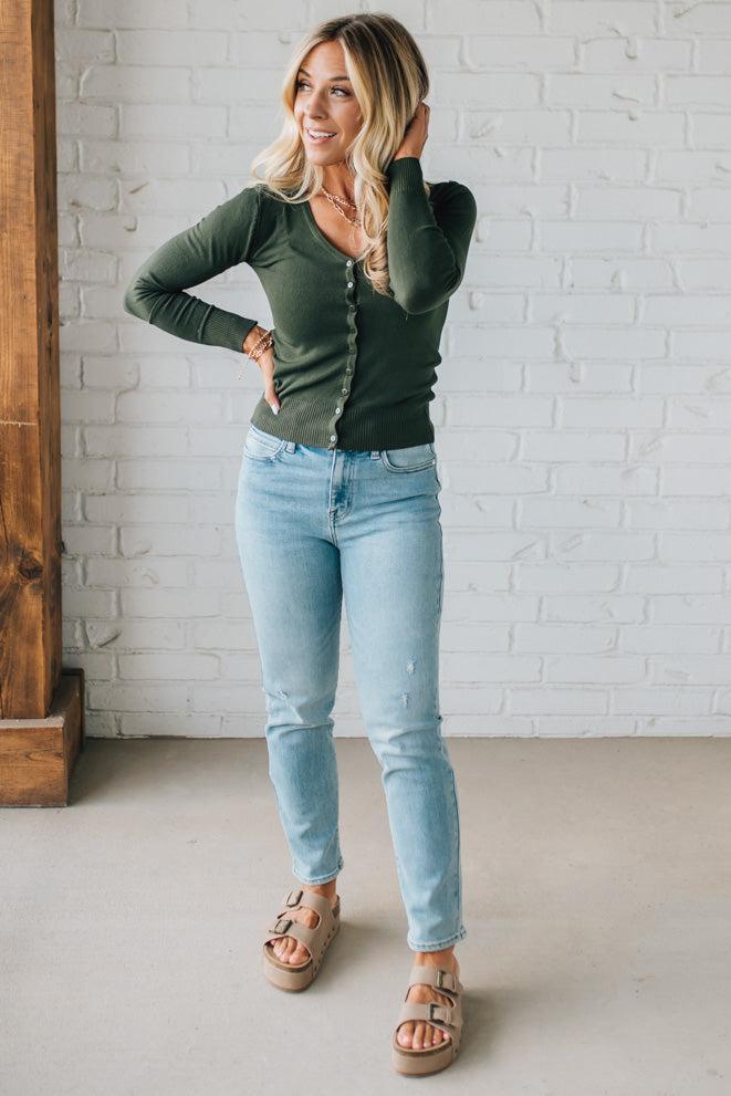 Woman wearing a green long-sleeve top and light blue jeans standing against a white brick wall.