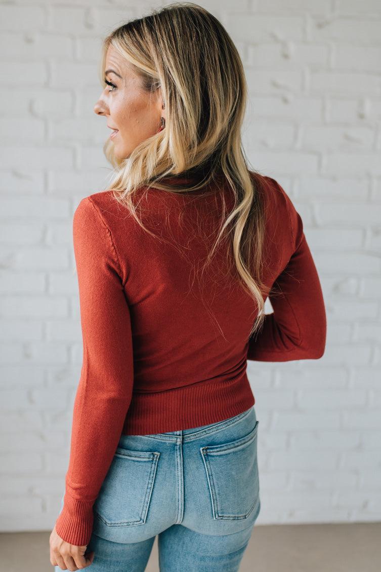 Woman wearing a red sweater and blue jeans against a white brick wall.