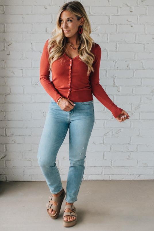 Woman wearing a red long-sleeve top and light blue jeans against a white brick wall.