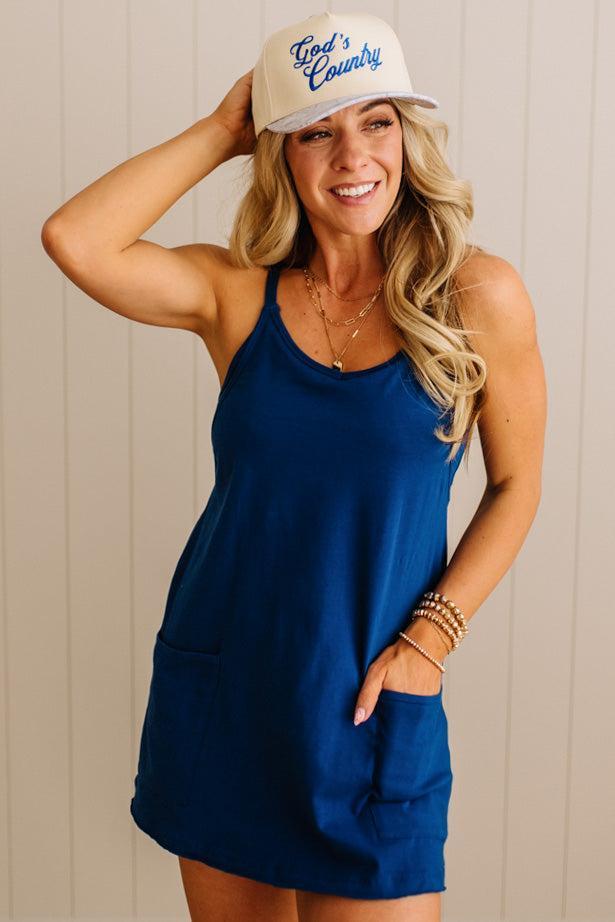 Woman wearing a blue dress and white cap with 'God's Country' text against a beige wall.