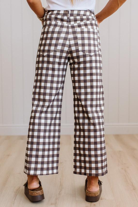 A person wearing brown and white gingham wide-leg cropped pants, paired with brown slide sandals and a white top, standing in a room with light wood floors and white paneled walls.