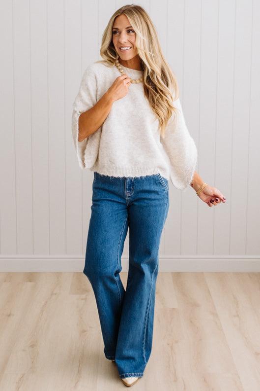 BLONDE WOMAN WEARING DARK JEANS IN A BRIGHTLY LIT STUDIO WITH A SOLID SWEATER FEATURING SCALLOP TRIM AND BUTTONS UP THE SLEEVE IN THE COLOR HEATHERED OATMEAL.