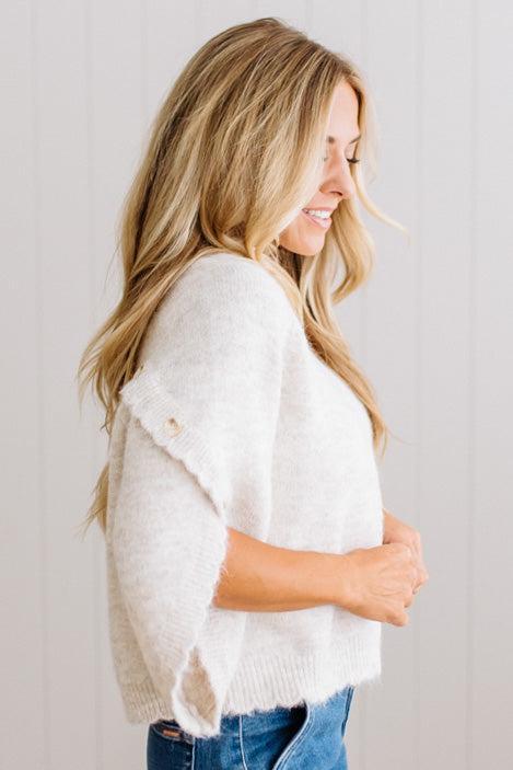 BLONDE WOMAN WEARING DARK JEANS IN A BRIGHTLY LIT STUDIO WITH A SOLID SWEATER FEATURING SCALLOP TRIM AND BUTTONS UP THE SLEEVE IN THE COLOR HEATHERED OATMEAL.