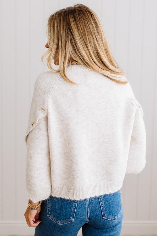 BLONDE WOMAN WEARING DARK JEANS IN A BRIGHTLY LIT STUDIO WITH A SOLID SWEATER FEATURING SCALLOP TRIM AND BUTTONS UP THE SLEEVE IN THE COLOR HEATHERED OATMEAL.
