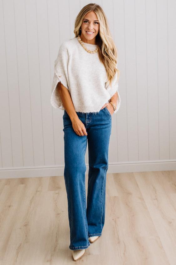 BLONDE WOMAN WEARING DARK JEANS IN A BRIGHTLY LIT STUDIO WITH A SOLID SWEATER FEATURING SCALLOP TRIM AND BUTTONS UP THE SLEEVE IN THE COLOR HEATHERED OATMEAL.