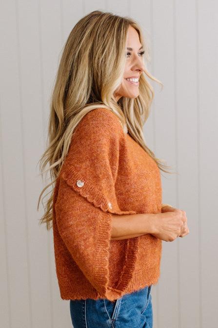 BLONDE WOMAN WEARING DARK JEANS IN A BRIGHTLY LIT STUDIO WITH A SOLID SWEATER FEATURING SCALLOP TRIM AND BUTTONS UP THE SLEEVE IN THE COLOR HEATHERED RUST.