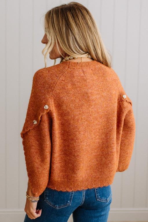 BLONDE WOMAN WEARING DARK JEANS IN A BRIGHTLY LIT STUDIO WITH A SOLID SWEATER FEATURING SCALLOP TRIM AND BUTTONS UP THE SLEEVE IN THE COLOR HEATHERED RUST.
