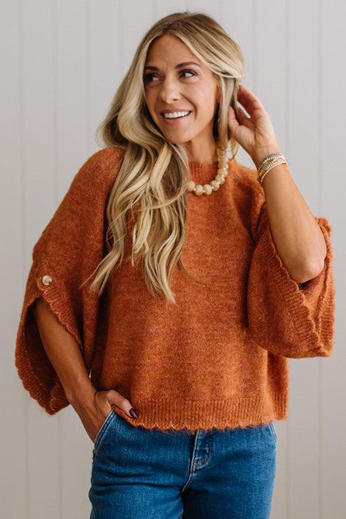 BLONDE WOMAN WEARING DARK JEANS IN A BRIGHTLY LIT STUDIO WITH A SOLID SWEATER FEATURING SCALLOP TRIM AND BUTTONS UP THE SLEEVE IN THE COLOR HEATHERED RUST.