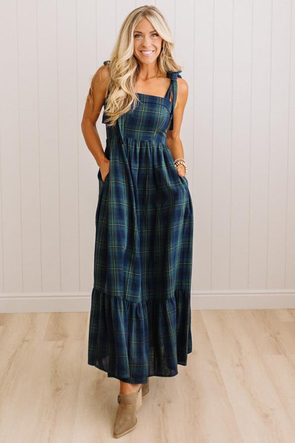 A woman modeling a sleeveless, tiered maxi dress with a dark blue and green plaid pattern, standing in front of a white paneled wall and light wood floor.