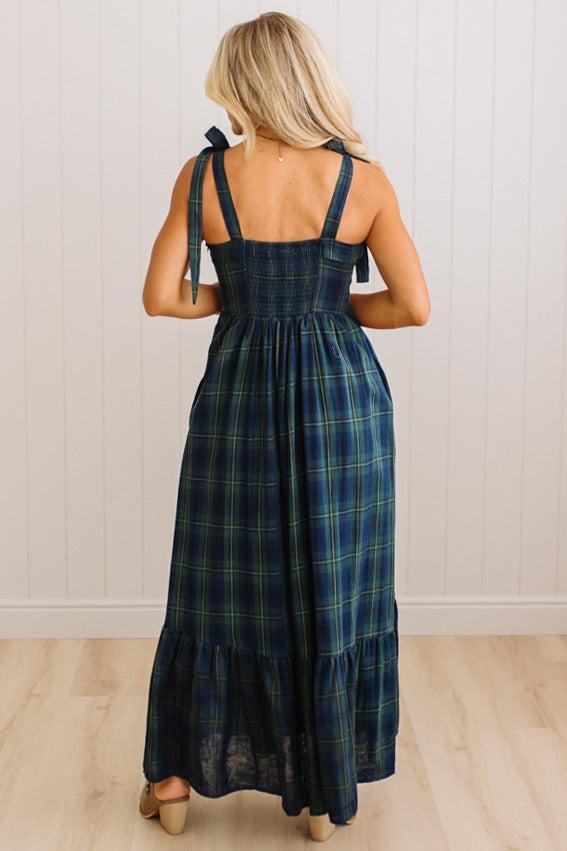 A woman viewed from behind wearing a dark navy blue plaid maxi dress with tie-shoulder straps, standing on a light wood floor against a white paneled wall.