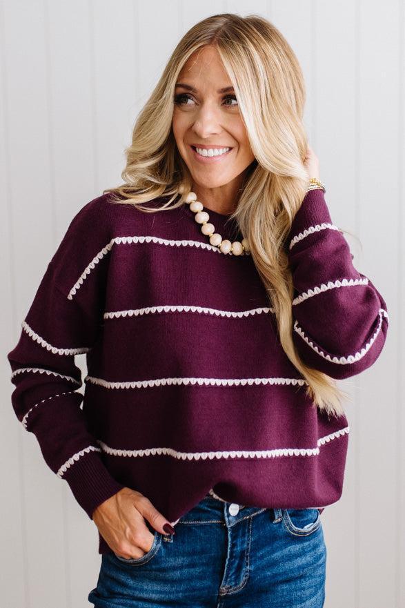 BLONDE WOMAN WEARING DARK JEANS IN A BRIGHTLY LIT STUDIO MODELING A CREW NECK, LONG SLEEVE, BLANKET STITCH STRIPED SWEATER IN DARK PLUM.