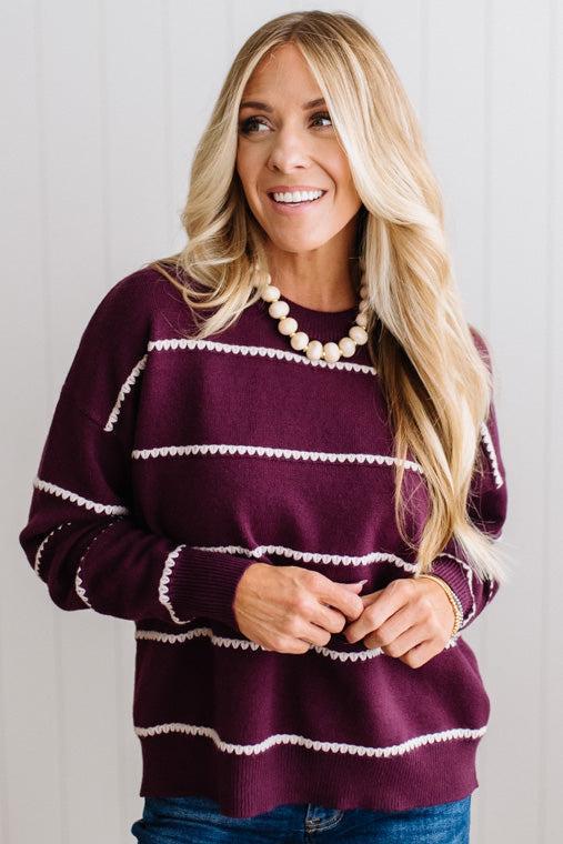 BLONDE WOMAN WEARING DARK JEANS IN A BRIGHTLY LIT STUDIO MODELING A CREW NECK, LONG SLEEVE, BLANKET STITCH STRIPED SWEATER IN DARK PLUM.