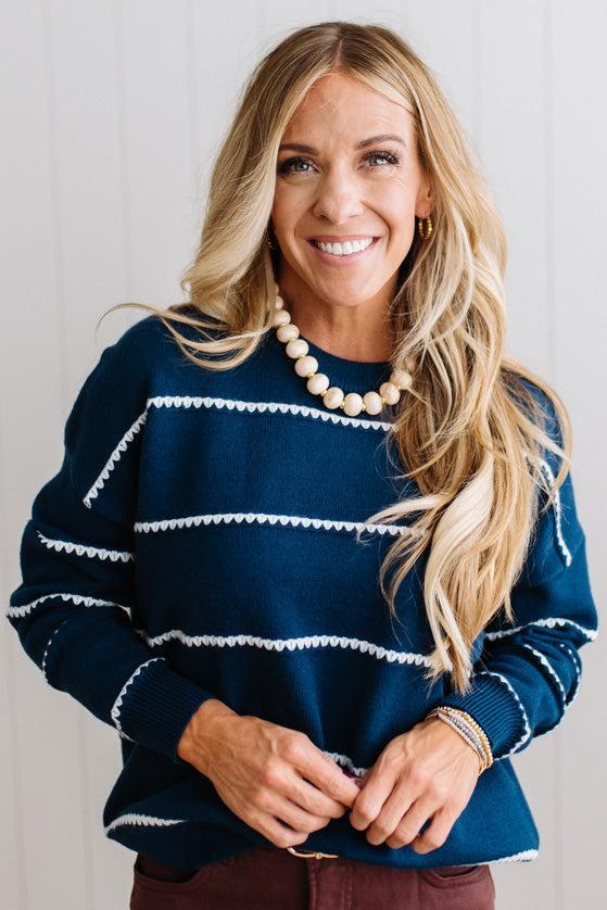 BLONDE WOMAN WEARING BROWN JEANS IN A BRIGHTLY LIT STUDIO MODELING A CREW NECK, LONG SLEEVE, BLANKET STITCH STRIPED SWEATER IN DARK TEAL.