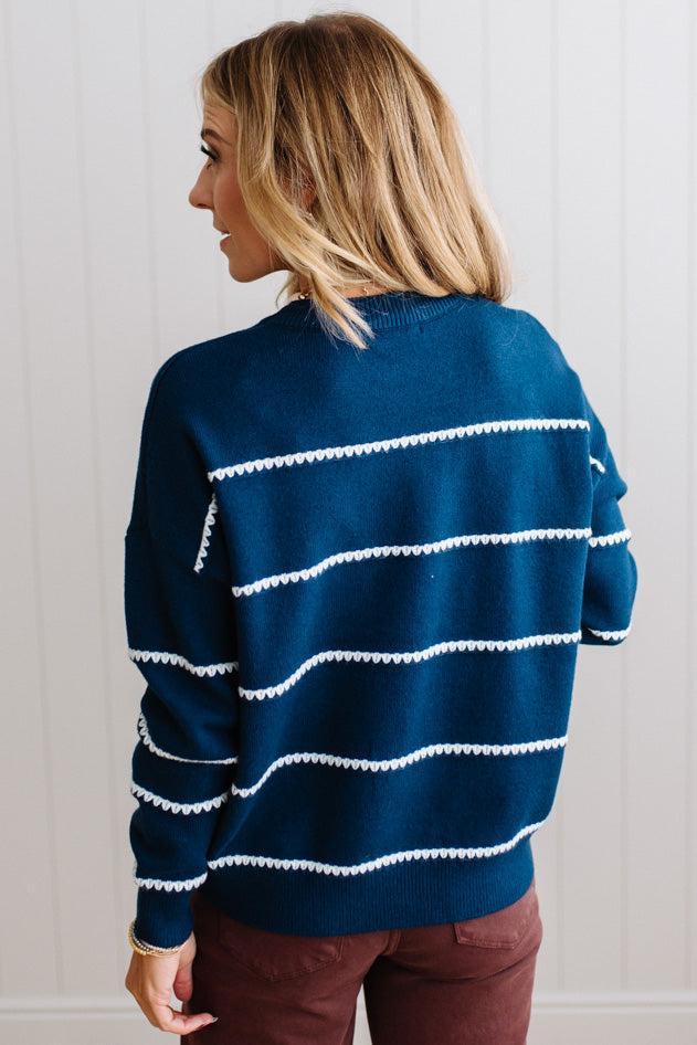 BLONDE WOMAN WEARING BROWN JEANS IN A BRIGHTLY LIT STUDIO MODELING A CREW NECK, LONG SLEEVE, BLANKET STITCH STRIPED SWEATER IN DARK TEAL.