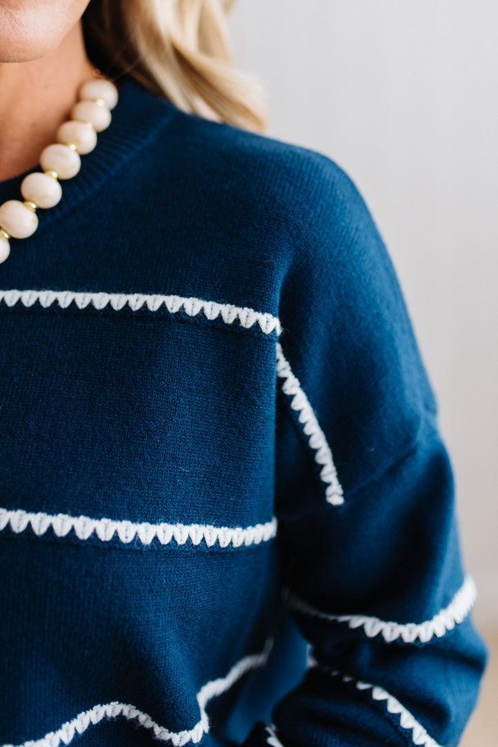 BLONDE WOMAN WEARING BROWN JEANS IN A BRIGHTLY LIT STUDIO MODELING A CREW NECK, LONG SLEEVE, BLANKET STITCH STRIPED SWEATER IN DARK TEAL.
