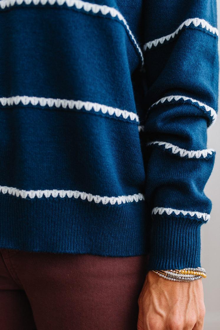BLONDE WOMAN WEARING BROWN JEANS IN A BRIGHTLY LIT STUDIO MODELING A CREW NECK, LONG SLEEVE, BLANKET STITCH STRIPED SWEATER IN DARK TEAL.