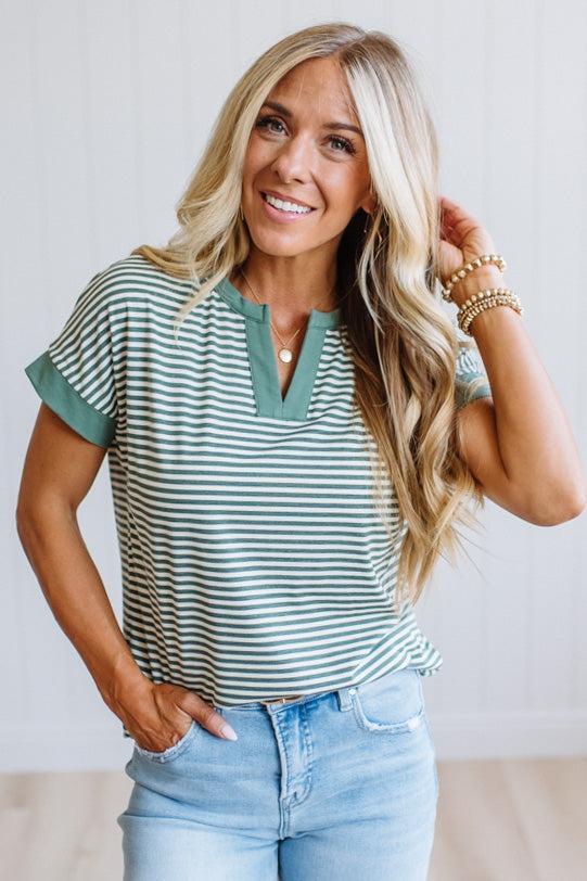 Woman wearing a green striped shirt and light jeans standing in a room with light wood flooring and white walls.