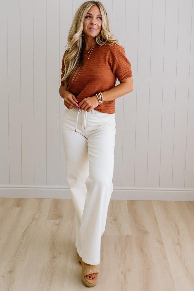Woman wearing a brown ribbed sweater and white pants standing against a light wooden paneled wall.