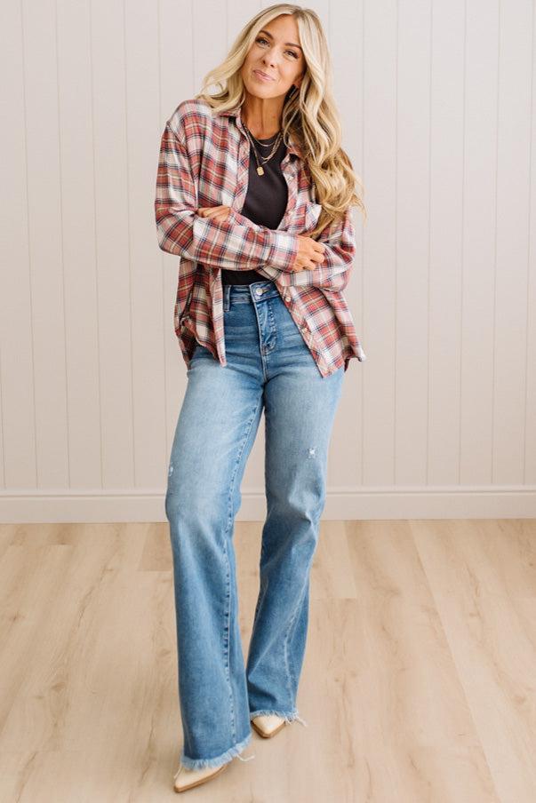A woman modeling a red and white plaid button-down shirt over a black top, paired with light-wash blue flare jeans and beige pointed-toe flats, standing in a room with light wood floors and white paneled walls.