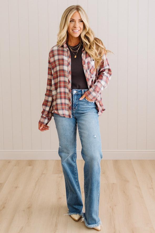 A woman modeling a red and brown plaid button-down shirt over a black top, paired with light-wash blue flare jeans, standing in front of a white paneled wall and light wood floor.
