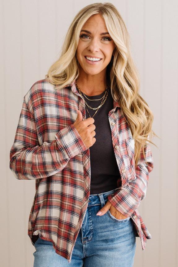 A smiling blonde woman wearing a red, white, and black plaid button-up flannel shirt over a black top and blue jeans, standing against a light beige paneled wall.