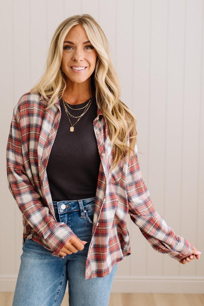 A woman modeling a red, white, and blue plaid button-down flannel shirt over a black top and blue jeans, standing against a light beige paneled wall.