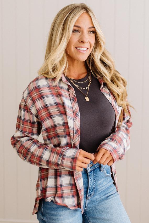 A woman wearing a red, white, and blue plaid button-up flannel shirt over a dark gray top and blue jeans, standing against a light beige paneled wall.