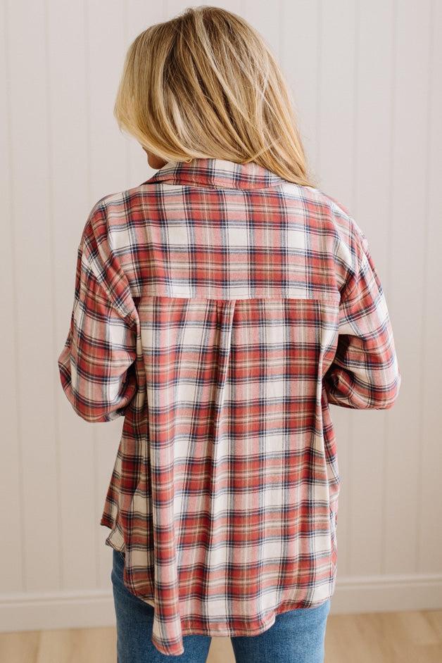 A woman viewed from behind wearing a long-sleeve plaid button-up shirt in red, white, and navy, paired with blue jeans, set against a light wood-paneled wall.